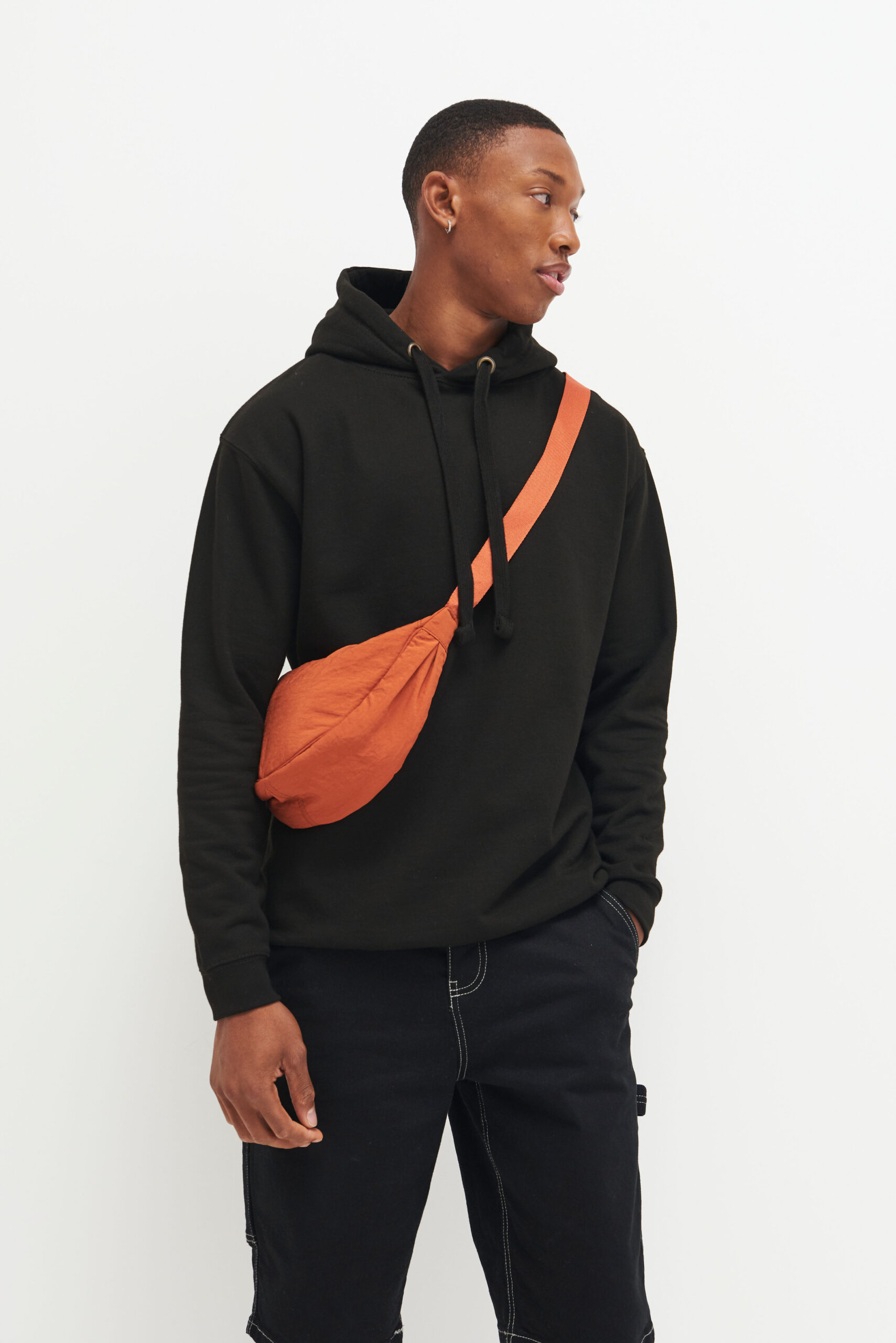 Model looking away wearing all black hoodie and an orange cross body bag.