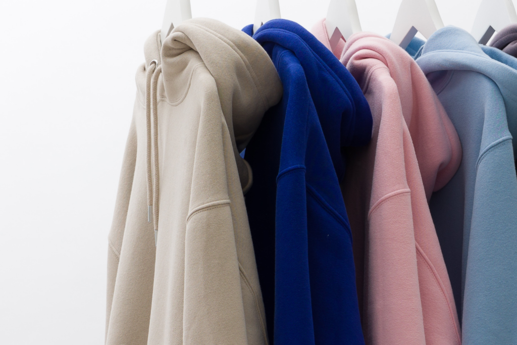 Five hoodies in beige, blue, pink, and pastel shades hanging on white hangers.