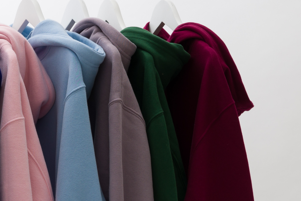 Five hoodies in pink, blue, grey, green, and burgundy hanging on white hangers.